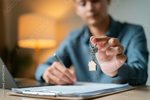 Person holds a house-shaped keychain while signing property agreement next to a miniature house model, laptop and clipboard on desk.