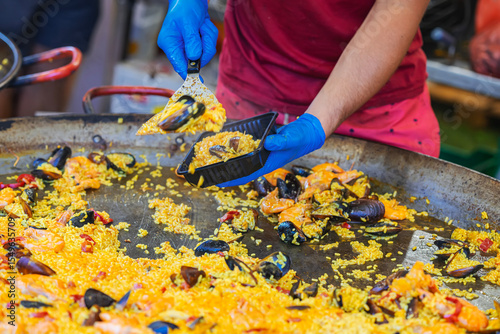 Chef in blue gloves serving seafood paella into plastic container from giant skillet during food event. Concept of paella, street food service. Spanish Culture and Food Festival