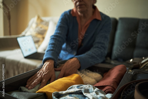 Canvas Print Elderly woman packing clothes into suitcase with open laptop on bed showing sere