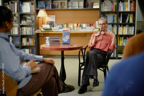 Senior Caucasian woman sitting on chair speaking into microphone gesturing with hand during session in bookstore with audience listening and books displayed on table