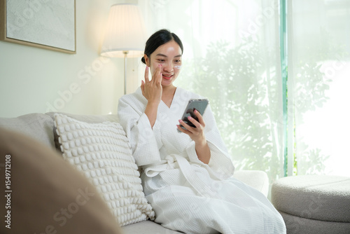 Asian female in bathrobe doing skincare and checking updates on smartphone at home. Self care concept.