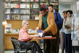 Caucasian woman sitting at table signing book for Black man while diverse group of people standing in line holding books in bookstore