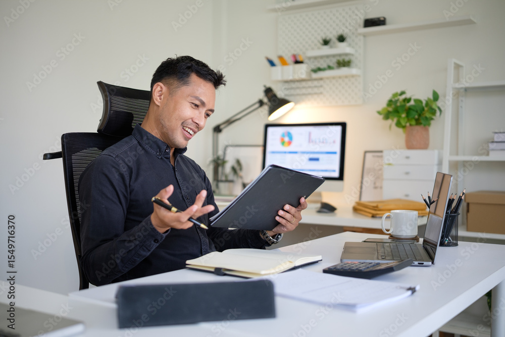 Naklejka premium Smiling asian businessman evaluating business reports at a modern desk.