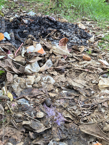 Charred fire pit with smoldering ashes and burnt leaf debris on dusty ground after garbage burning session