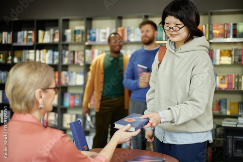 Asian young adult woman receiving book with autograph from Caucasian female author during session, multiethnic group of young adult men standing in background holding books