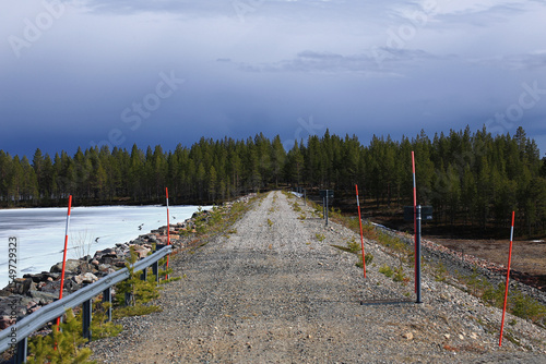 Gravel road on dam with ice-covered reservoir and red snow poles under a dark blue sky