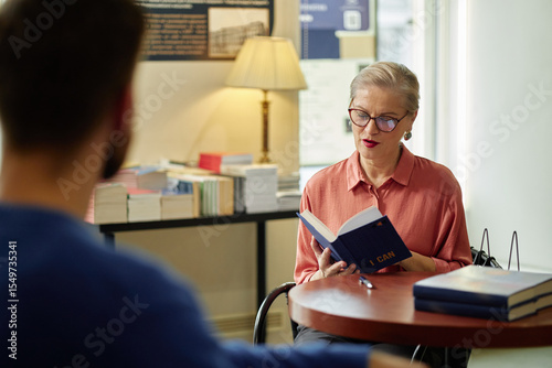 Obraz na plátne Caucasian senior woman wearing glasses reading book aloud at round table in libr