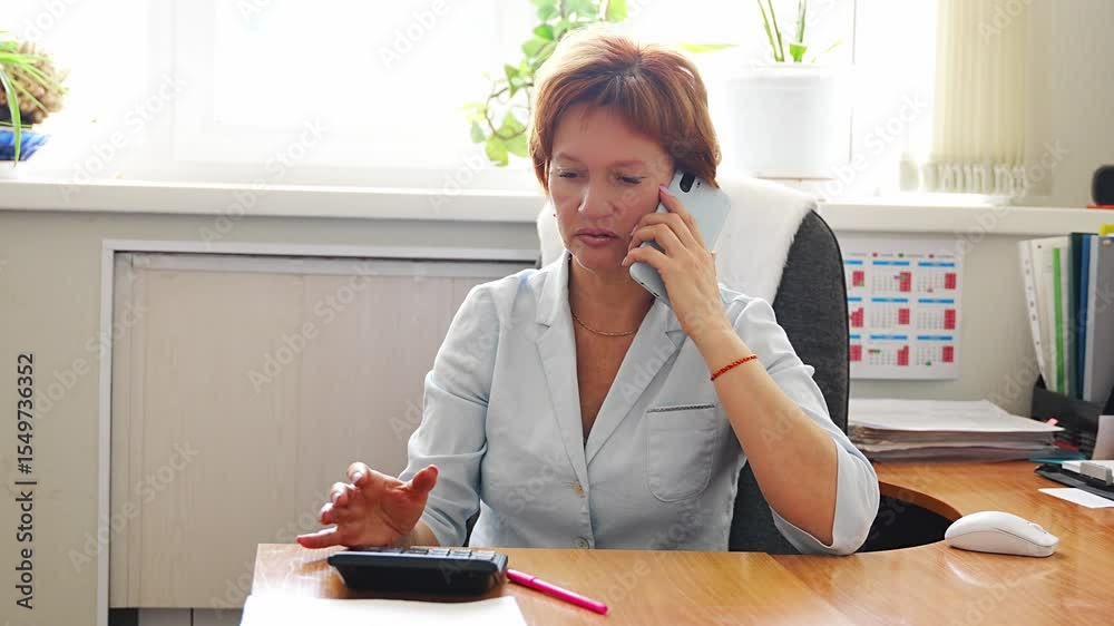 Woman sitting at her desk in the office, talking on the phone while working on the computer. Business communication, multitasking, and everyday coordination at the workplace.