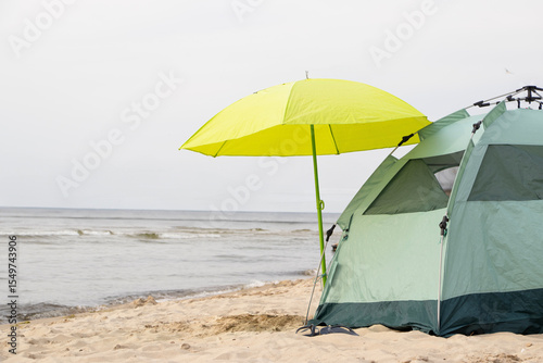 A green tent with a yellow umbrella stands on a sandy beach by the sea. A peaceful seaside camping spot under the bright summer sun.
