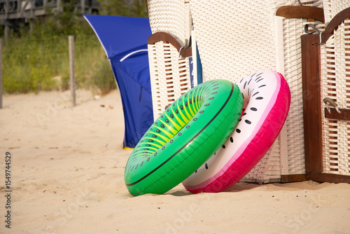Two inflatable swim rings, one green and one pink, stand upright on a sandy beach near the sea, ready for summer fun and relaxation.