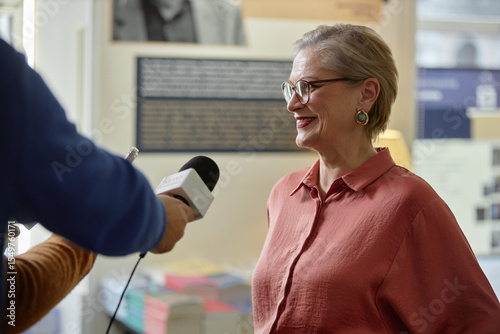 Caucasian middle aged woman smiling while being interviewed by unseen holding microphone, standing indoors with blurred books and informational displays in background