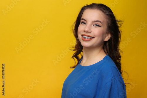 Positive teen girl with dental braces in blue t-shirt. Hands behind head, wide smile, looks at camera on yellow backdrop. Dental care services, orthodontic clinics, teen health campaigns, youth