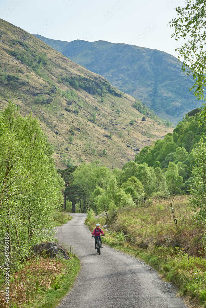 active senior woman riding her electric mountain bike in the Glen Nevis valley below Ben Nevis summit in the highlands near Fort William in Scotland, UK active senior woman riding her electric mountain bike in the Glen Nevis valley below Ben Nevis summit in the highlands near Fort William in Scotland, UK