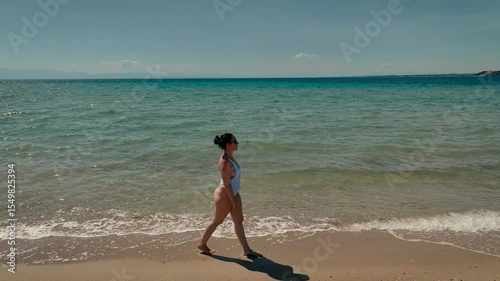 Woman in a striped swimsuit and sunglasses walking along the shoreline of a beautiful sandy beach, with gentle waves from the turquoise sea.
