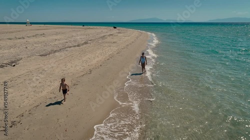 Aerial view of a happy mother and her son running and playing on a beautiful, wide sandy beach with turquoise water during a sunny summer family vacation.