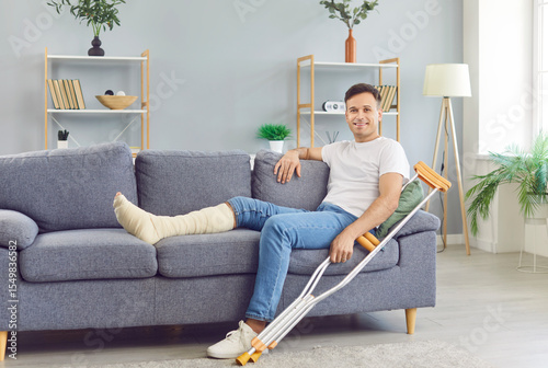 Tableau sur toile Young smiling man with crutches sitting on sofa at home on rehabilitation with leg in plaster cast after injury