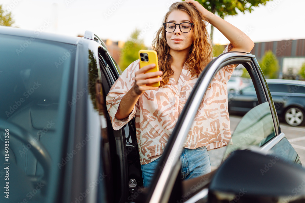 Naklejka premium Beautiful woman with curly hair with a phone in her hands stands near a car in a parking lot. Young female driver enjoys the weather and chats at sunset. Transport and technology concept.
