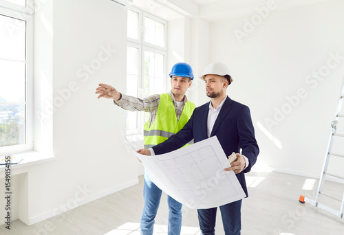 Two men in helmets with a drawing of the building inspect the empty room. The foreman shows the work done according to the plan. The contractor checks the work. Concept construction