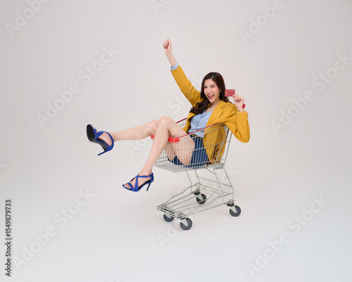 Ecstatic Asian woman in yellow blazer and heels holding a credit card joyfully sits in a shopping cart. Perfect for sales, promotions, or payment themes.
