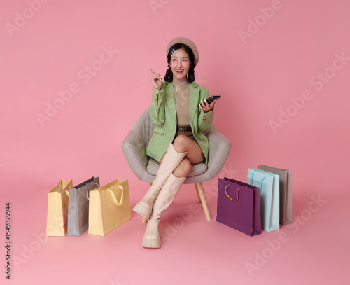 Joyful Asian woman in stylish outfit with beret relaxes in a chair holding mobile phone in hands shopping online isolated on pink background.