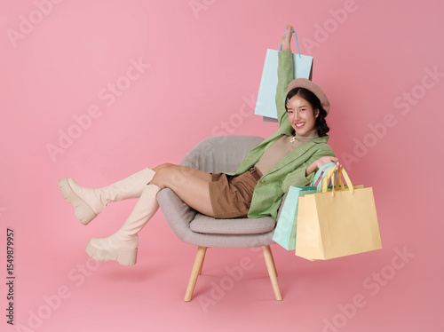 Joyful Asian woman in stylish outfit with beret relaxes in a chair holding up colorful shopping bags isolated on pink background