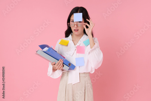 Asian woman feeling stressed with work overload, covered in colorful sticky notes and holding folders. Studio portrait on pink background, representing office pressure and multitasking.