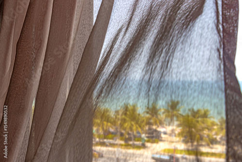 Soft transparent curtains blowing in a window with a tropical view of the sea beach with palm trees
