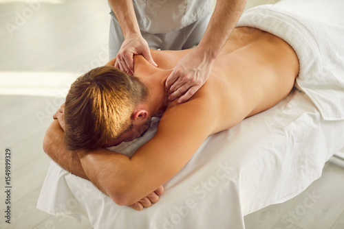Young man client lying on massage table receiving remedial body back and shoulder massage from professional masseur or doctor physiotherapist in clinic. Physiotherapy and rehabilitation concept.
