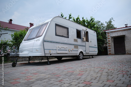 A white camper trailer is parked on a paved backyard near a brick house and cherry tree. RV travel content, camping promotions, suburban lifestyle visuals, or articles about alternative housing