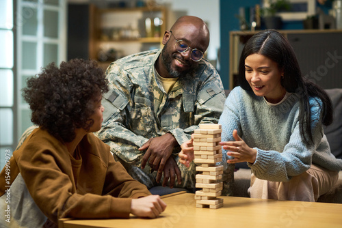 Caucasian woman and child playing wooden block game with smiling Black middle aged man in military uniform watching at home, family spending quality time together in living room