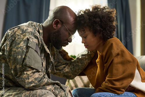 Black middle aged man in military uniform comforting Black young adult woman at home, both sitting close with foreheads touching, showing emotional family support and connection