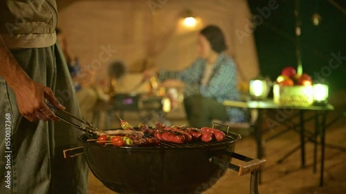 Man grilling sausages at campsite with his friend. Group of teenagers prepare dinner at campsite meal with grilled food and cozy lighting. Enjoying night picnic and barbecue in natural breeze.