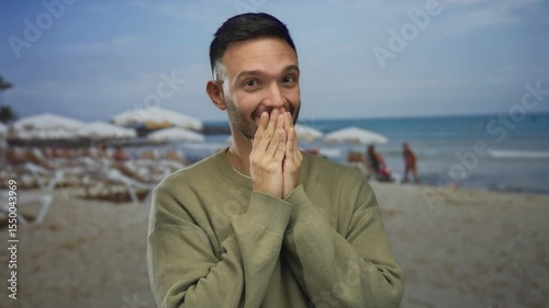 Young man with surprised expression covers mouth with hands on a sandy beach with ocean and umbrellas in the background, capturing an unexpected moment during a sunny seaside day.