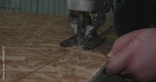 A professional electrician carefully cuts OSB panels with a precision jigsaw, following a precise line and producing shavings in the workshop. A man cuts a piece of OSB sheet with a jigsaw.