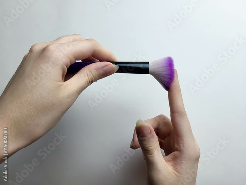 The hands hold a black cosmetic brush with lilac bristles