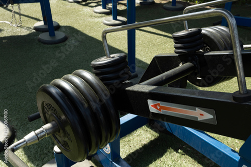 A close-up of a barbell with heavy weights on a gym bench. The setting is an outdoor gym with green turf flooring and blue equipment.