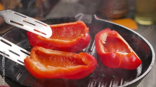 Closeup shot of kitchen tongs turning red bell pepper slices on a hot grill pan with visible smoke rising. The searing process chars the vegetable for a delicious and savory appetizer