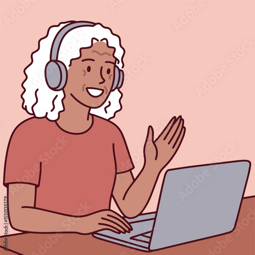 Senior Dark skinned woman with curly hair wears headphones and waves while communicating remotely via laptop in online meeting