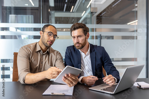 Two businessmen reviewing a tablet and laptop, discussing business strategies in a modern office setting.