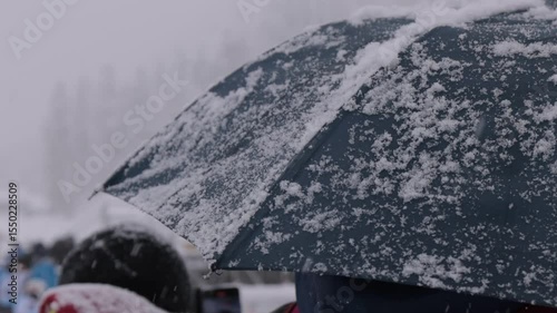 The image shows a snow-covered umbrella with a gray fabric, set against a snowy landscape with pine trees in the background. A person in a red and black jacket stands nearby, facing the scene under a