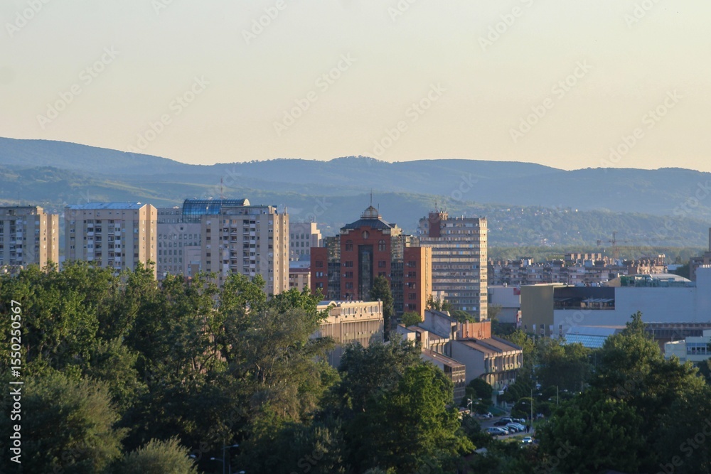 Obraz premium Panoramic view of Novi Sad, Serbia, featuring modern residential buildings, lush greenery, and distant mountains under soft evening light.