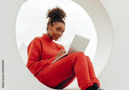 Woman in red outfit using laptop and phone, sitting in a circle looking at camera.
