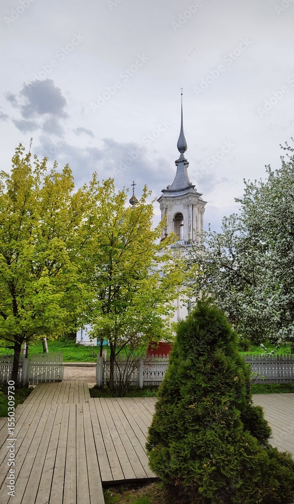 Naklejka premium Church of Varlaam of Khutynsky Ilyinsky parish surrounded by flowering trees. Bell tower of ancient Orthodox Church and spring greenery. Russia, Vologda, May 23, 2025