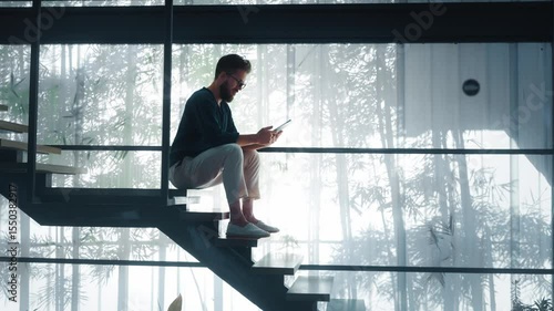 Creative professional sitting on stairs in modern office space, using tablet while enjoying natural light streaming in. Creative professional billionaire who created successful project