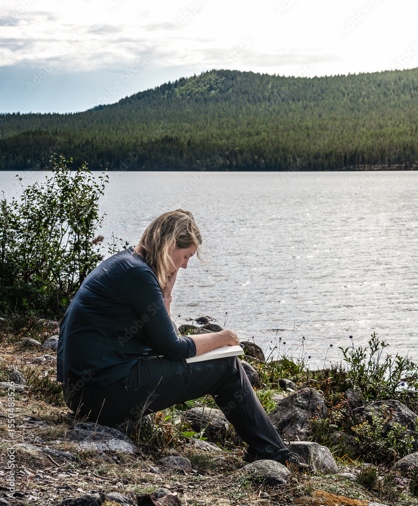 Fototapeta premium Outdoor woman reading a book while sitting at the shore of a lake on a bright summer day in Sweden. She is enjoying the peace and quiet of nature.