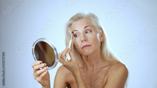 A mature woman with long hair smiles happily as she applies makeup in a serene spa setting. The calming background enhances her joyful experience.