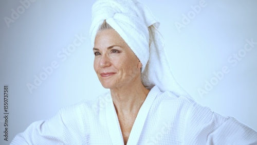 A mature woman smiles joyfully while pampering herself at a spa. She wears a white robe and towel, radiating relaxation and happiness against a light background.