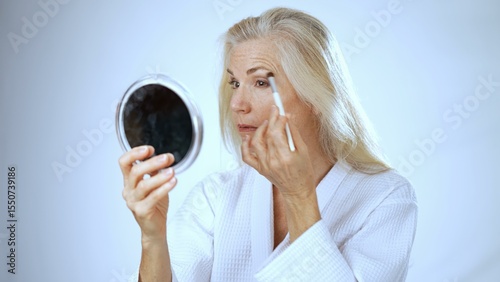 A mature woman smiles contently in a spa setting, applying makeup with a brush. The serene white background enhances her joyful expression and self-care moment.