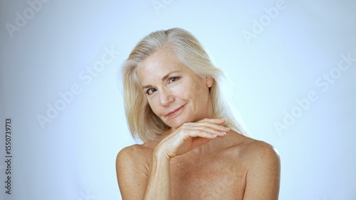 A mature woman with long, light hair smiles happily while resting in a serene spa environment. The bright white background enhances her joyful expression.