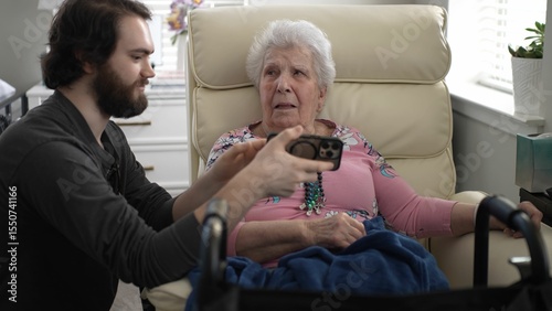 A young man shares images on his smartphone with his grandmother, sitting in a cozy chair. Their expressions reflect happiness and connection in a warm, home setting.